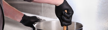 person wearing black nitrile gloves and stirring a large pot